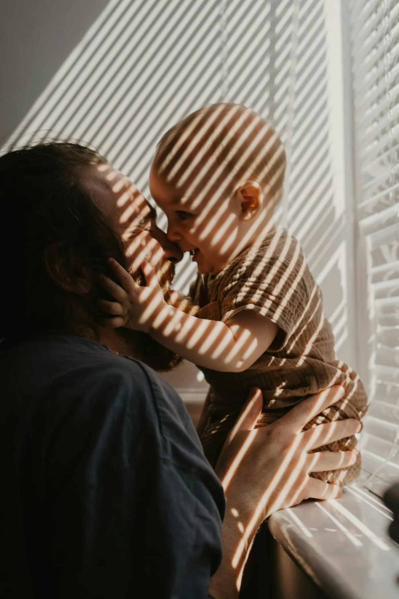 Padre y bebé junto a la ventana, las sombras de la persiana atraviesan sus rostros